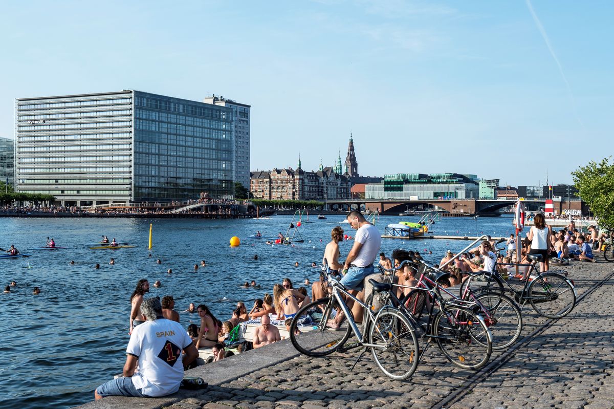 A group of people sitting along the waterfront of a city. Some people are swimming in the water.
