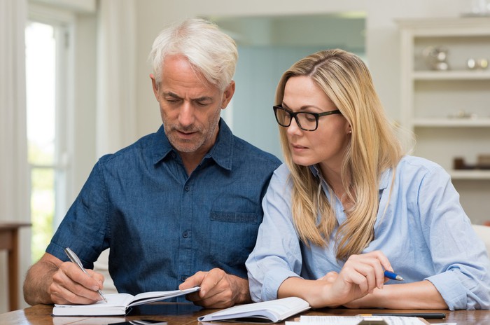 Older man writing in notebook while woman looks on.