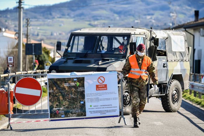 A soldier wearing a mask lifts up a barrier in front of an army truck with a rugged hill in background.
