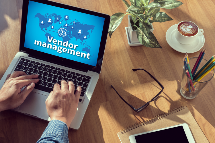 Man's hand behind computer showing Vendor Management; on table is coffee, plan, glasses, pencils, etc.