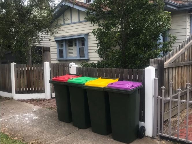 Four bins with red, green, yellow and purple lids sit in a row on the footpath outside a house.