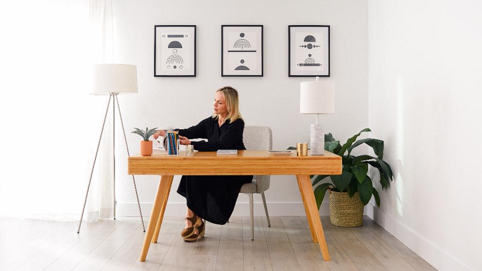 A woman sitting at a wooden desk rented from Fernish, with lamp[, a chair, and wall art, also from Fernish, a furniture subscription rental service.