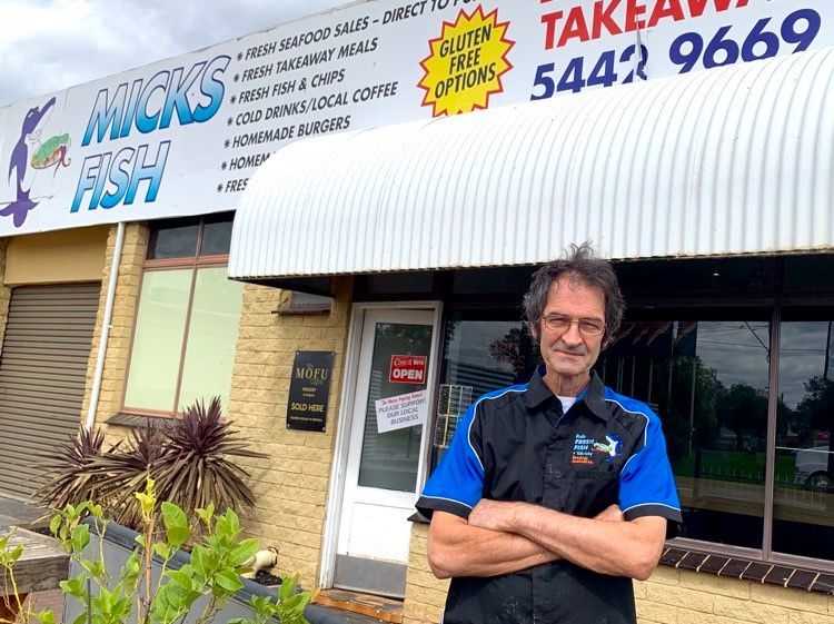 A man is standing outside a fish and chip shop folding his arms, wearing glasses looking at the camera.