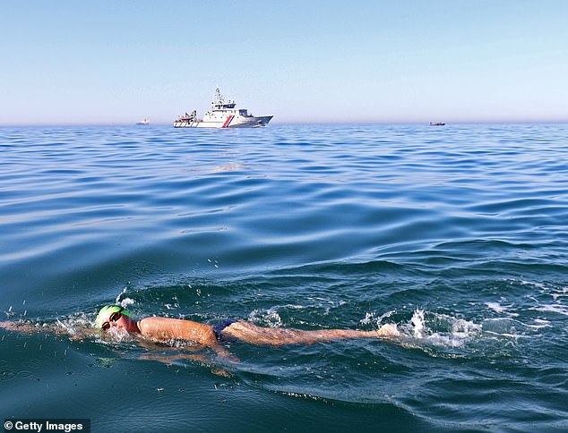 Pictured: A swimmer crosses the English Channel as, in the background, a dinghy carrying migrants is escorted by French border police on Friday