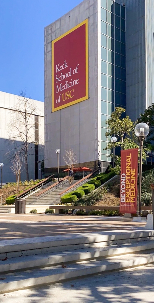 Photo of the Keck School of Medicine, a tall gray building with a banner reading “Keck School of Medicine of USC.” There are stairs leading up to the building, surrounding foliage, another building, and a blue sky in the background.