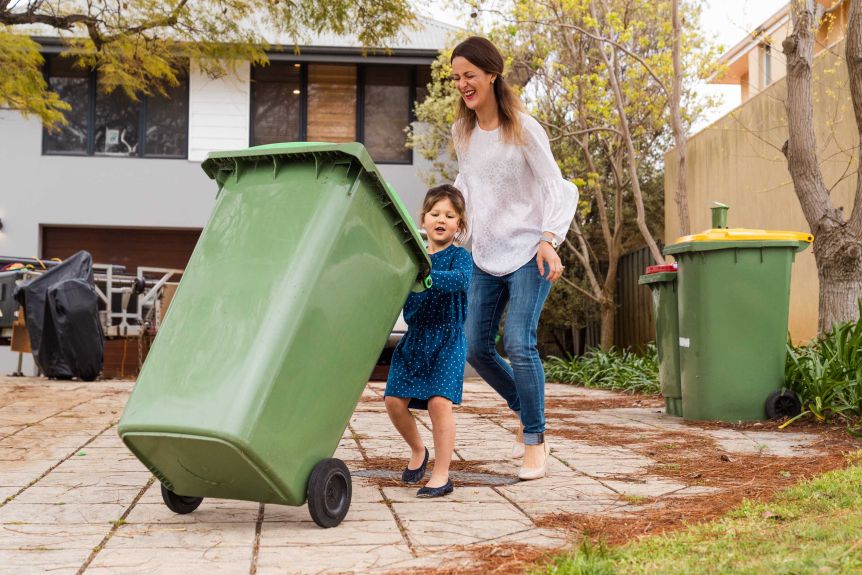 A small girl in a blue dress and a woman wearing jeans push a green bin on a driveway.