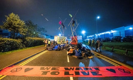 Scene of a road blocked by protesters and vans at night, with a large orange banner saying 'We Want To Live' spread out on the road.