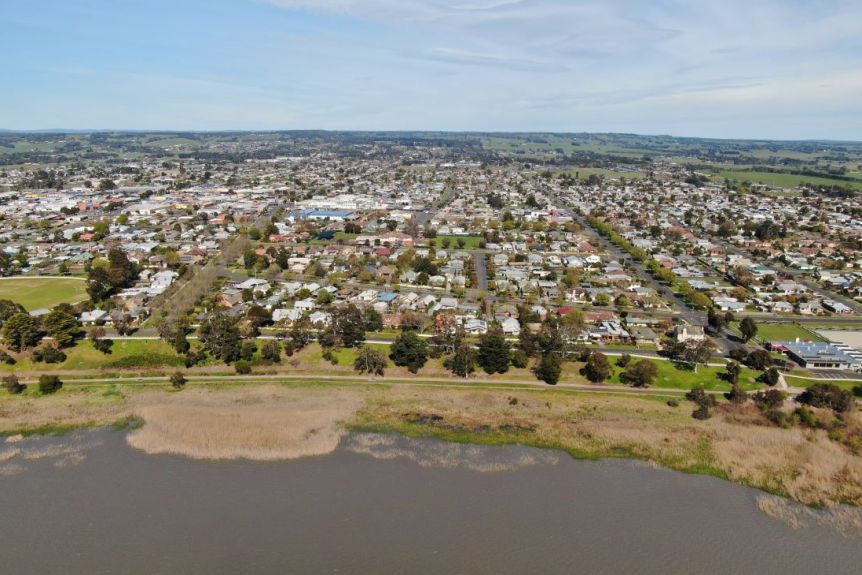 An aerial photo of a country town built on the edge of a lake, surrounded by green paddocks and hills.