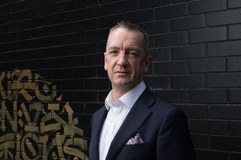A man, slightly frowning, in a black suit standing in front of a black brick wall.