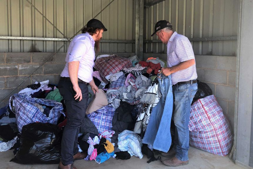 Two men standing in front of a large pile of clothing inside a corrugated iron shed.