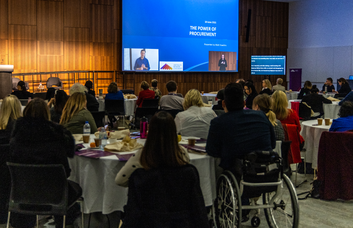 Crowd of people at AND's Conference looking at Matt Hawkins talking on the screen. Auslan Interpreter and captioning shown. PowerPoint slide saying 'The Power of Procurement'.