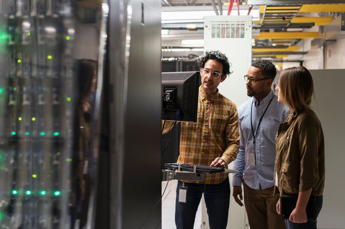 Three people work on a computer in a data center.