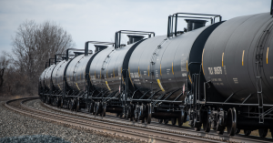 Several black rail tanker cars on a railway over gravel come around a corner in a forest of trees.