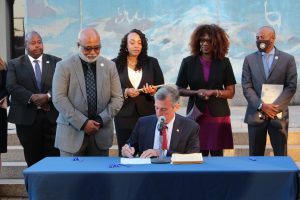 Governor Carney signs legislation while seated at a table as members of the General Assembly stand beside him.