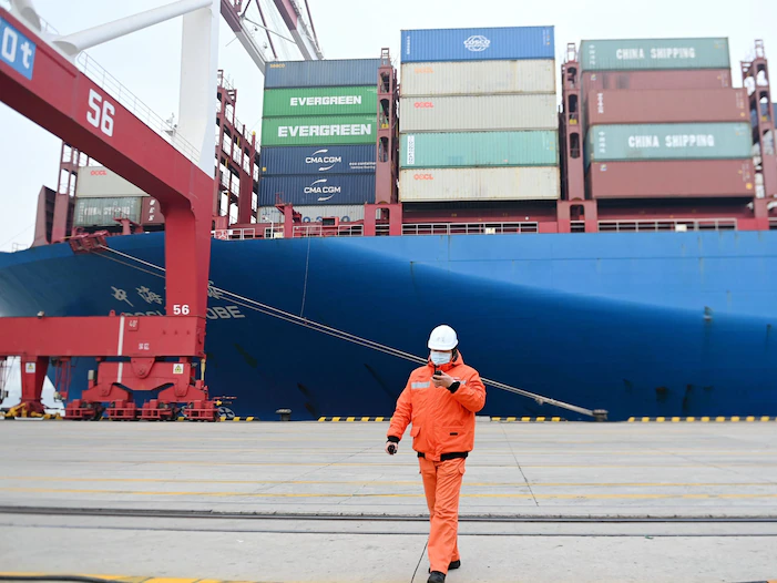 A masked man in orange in front of the side of a container ship in China.