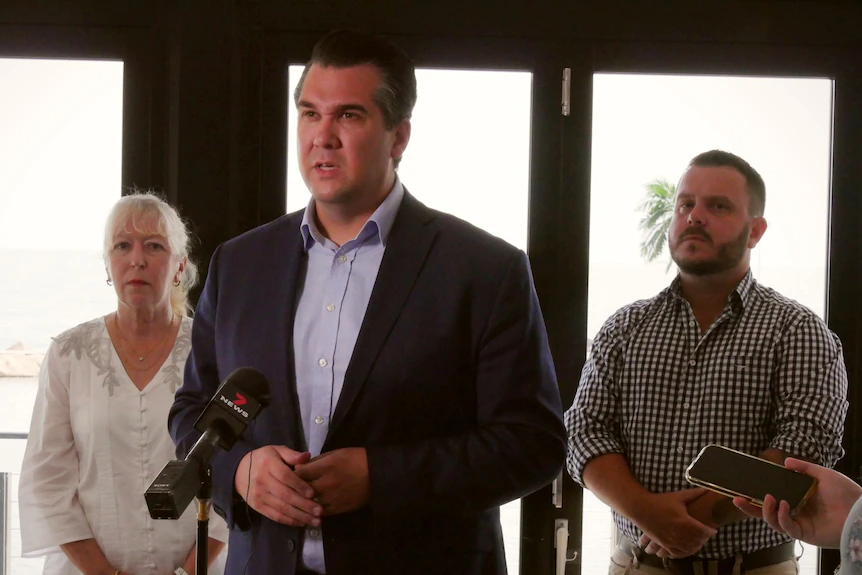 A man in a dark suit, flanked by a woman and another man, speaks to the media.