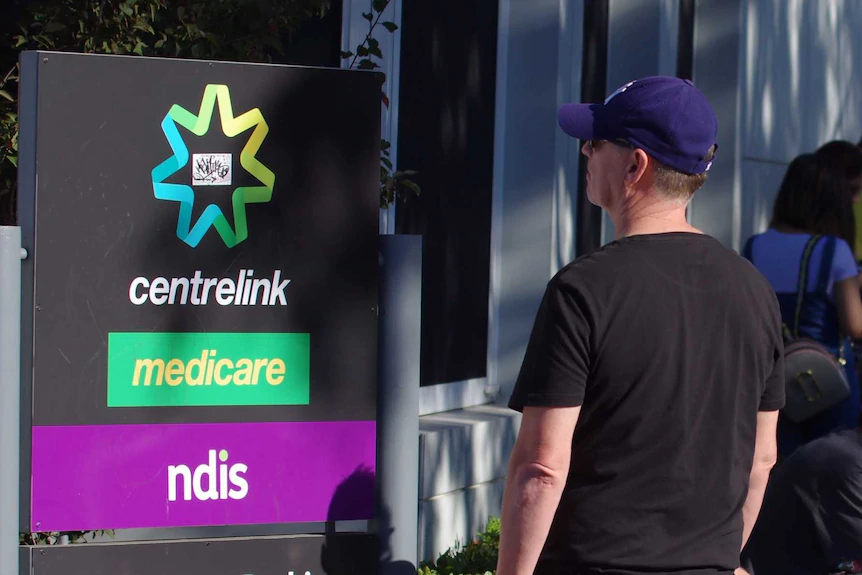 People line up outdoors beside a Centrelink sign.