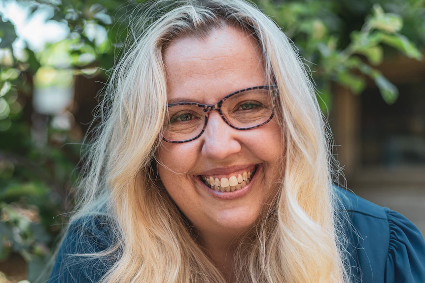 A woman with long blonde hair and brown rimmed glasses smiles for a photograph