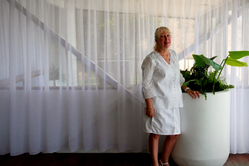 A woman in a white shirt and skirt with white-grey hair stands by a pot plant in a restaurant.