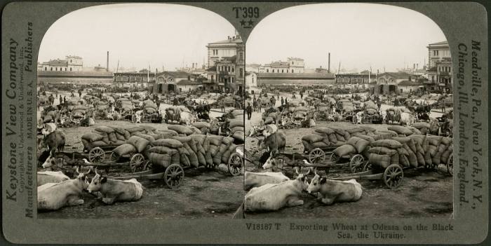 A black-and-white image of sacks of grain lie piled up near the harbourside buildings