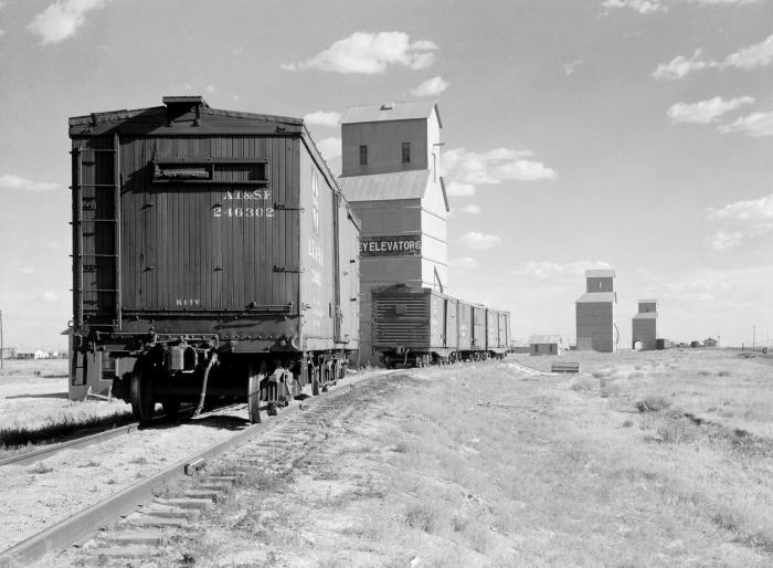 A black-and-white photo of freight train carriages on the approach to tall grain elevator structures