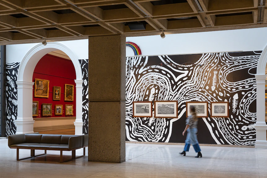 A gallery wall covered in a black and white linocut design, with black and white prints on it, and a stainglass rainbow.