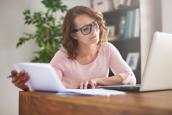 A person sitting at a desk and working on a laptop while holding papers in one hand.