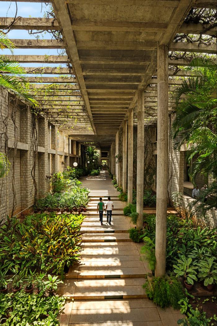 People walk along a colonnade which is partially open to the sky, in a modernist brick and concrete building with abundant plants