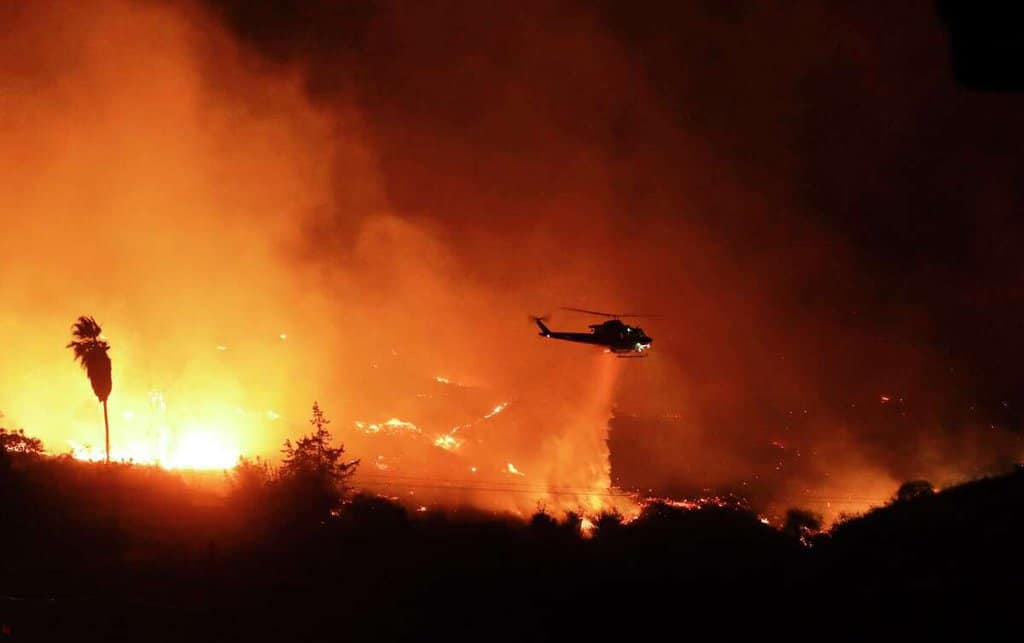 A helicopter douses a raging blaze in Bonsall, California (December 7, 2017). This wildfire event caused mandatory evacuations. Thus, environmental organizations argue in California courts that developers don't adequately evaluate wildfire hazards and choked evacuation routes when planning new developments close to fire-prone areas.