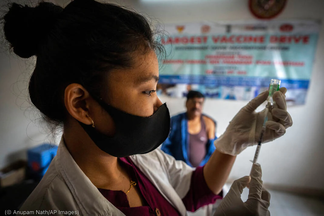 Woman wearing mask and filling syringe while patient waits in background (© Anupam Nath/AP Images) Woman wearing mask and filling syringe while patient waits in background (© Anupam Nath/AP Images)