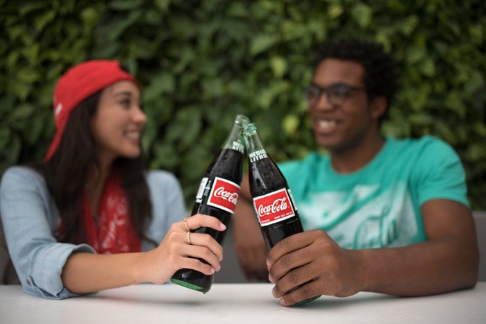Two people clinking their Coke bottles together while chatting and seated outside. 