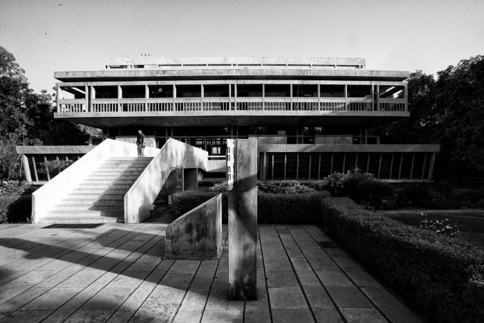 A low modernist concrete building is approached by a steps, surrounded by well-tended plants and trees