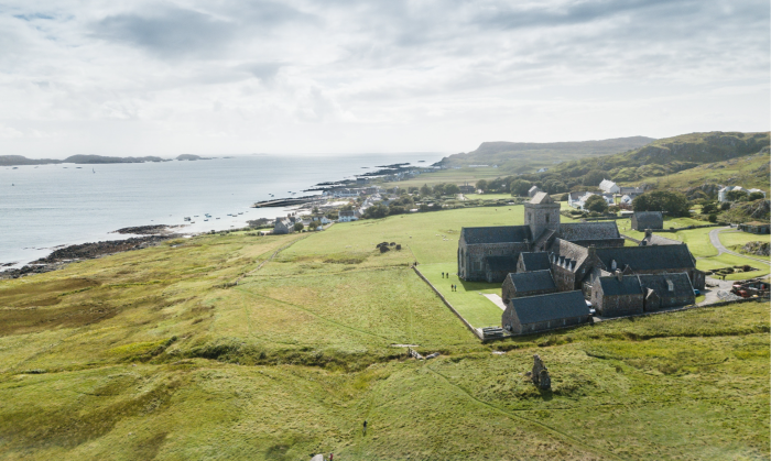 View of a stone abbey set among green moorland with houses beyond and the sea in the distance