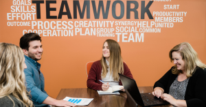 Four people sitting around a table discussing business topics underneath a sign that says Teamwork.