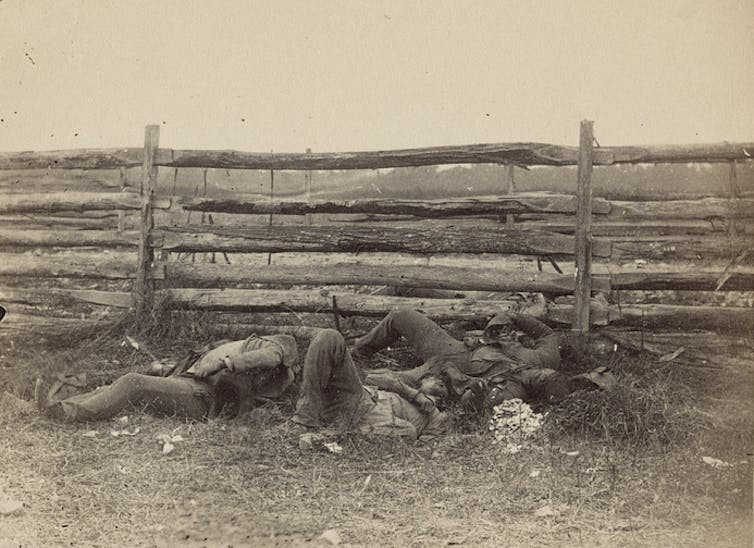 A black and white photograph of three dead bodies lying next to a split-rail fence.