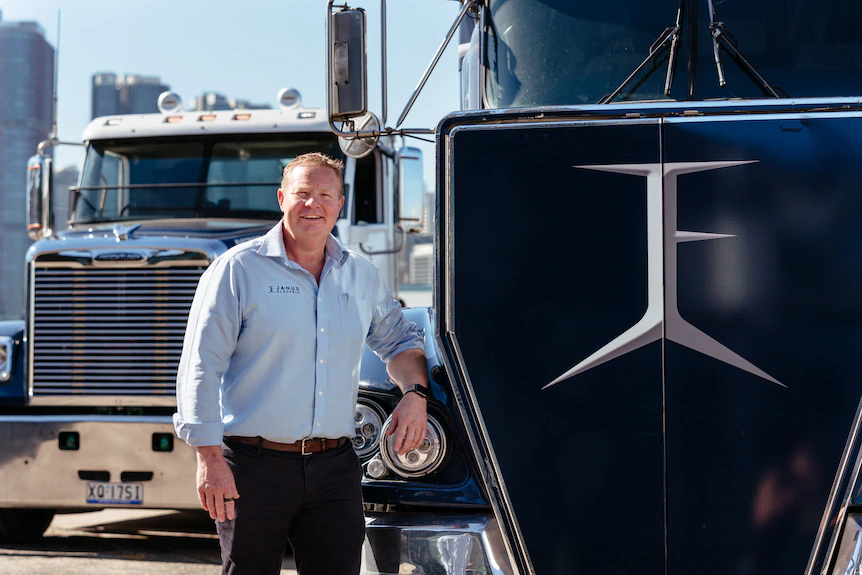 Man leaning on the fender of a truck with a second truck in the background.
