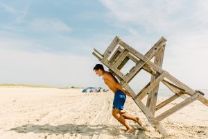 A lifeguard with the DNREC Division of Parks and Recreation Beach Patrol drags a lifeguard stand onto a sandy beach at the start of a sunny beach day.