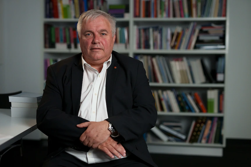 A man in white collared shirt wearing a black jacket sits looking at the camera in front of a book case
