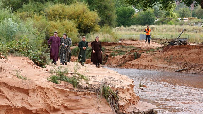 With a searcher in the background, local residents walk along the edge of Short Creek where two vans were swept away the day before on September 15, 2015 in Colorado City, Arizona. Flash floods from heavy rains on the afternoon of September 14, 2015 washed away two cars in Hildale as they were crossing a flooded creek, killing 13 people.