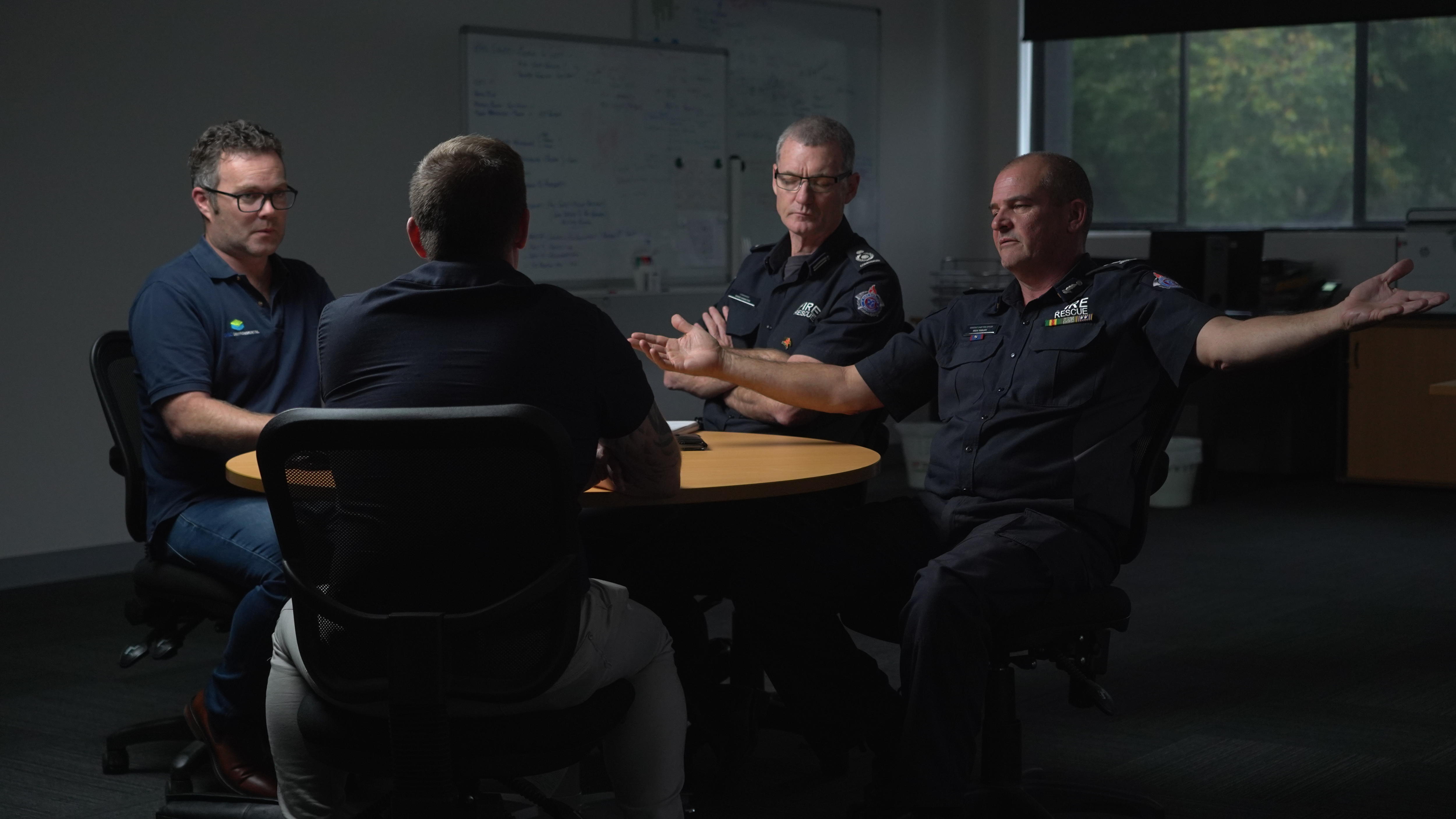 Four male firefighters in navy uniforms chat at a round table