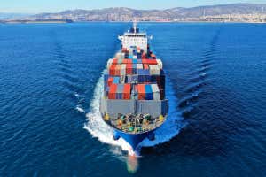 An above view of the front of a container ship moving through deep, blue ocean waters on a sunny day with dry hills in the background on shore. The ship is fully loaded with colourful, truck-sized cargo containers.