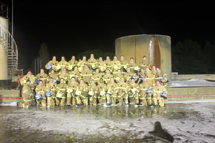 A group photo of 38 firefighters in yellow uniforms and holding helmets, stand for a group photo at training centre outdoors