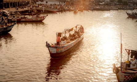 A traditional long wooden fishing boat motors through a calm harbour with the sun low in the sky making the water seem silvery