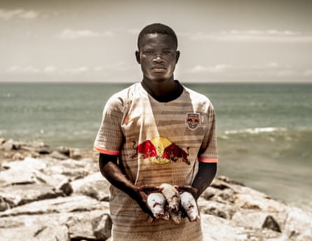 A young man by the sea in an old football shirt holding some fish