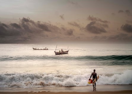 A man stands on the beach with a wooden board under his arm, a plastic bucket and a jerrycan